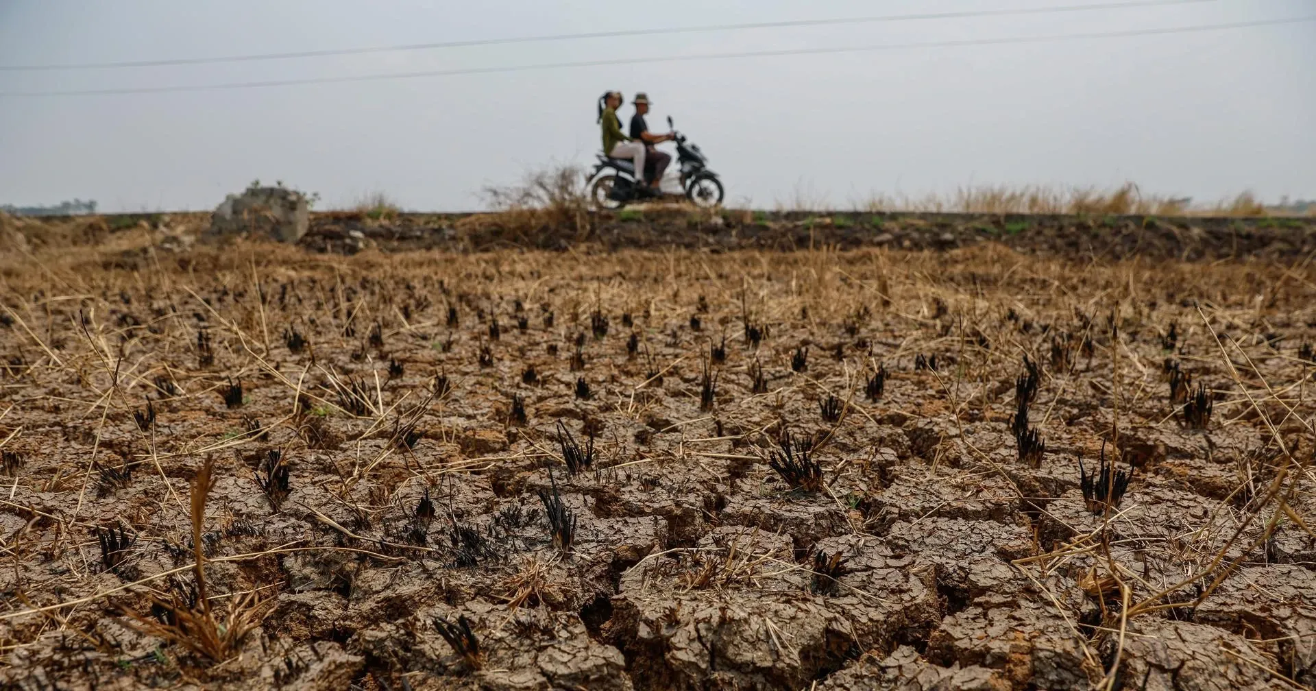 Tierra agrietada por sequía con moto de dos personas al fondo.