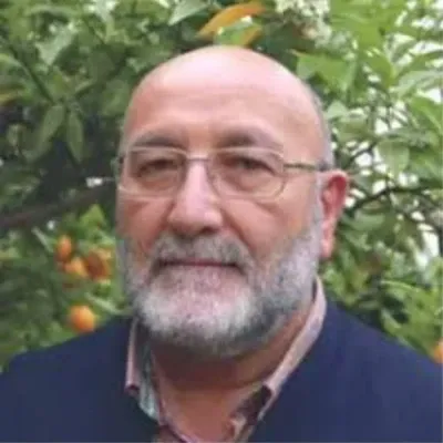 Alfonso Lucas, con gafas de montura fina, barba blanca y jersey azul marino, ante un fondo de vegetación.
