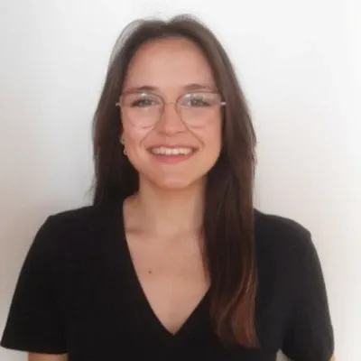 Carmen María Lais Gallardo, con camiseta negra, gafas de montura fina y cabello largo liso, sonriendo, retrato en primer plano sobre fondo blanco.
