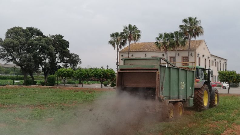Tractor con remolque esparciendo material en un campo verde con polvo visible.