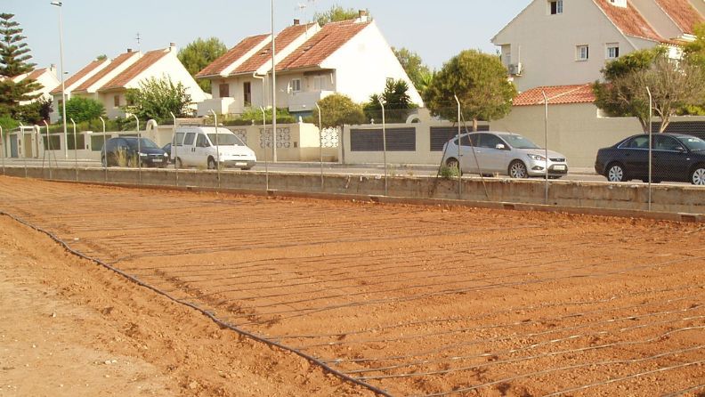 Terreno baldío con varillas de hierro, al fondo autos estacionados y casas.