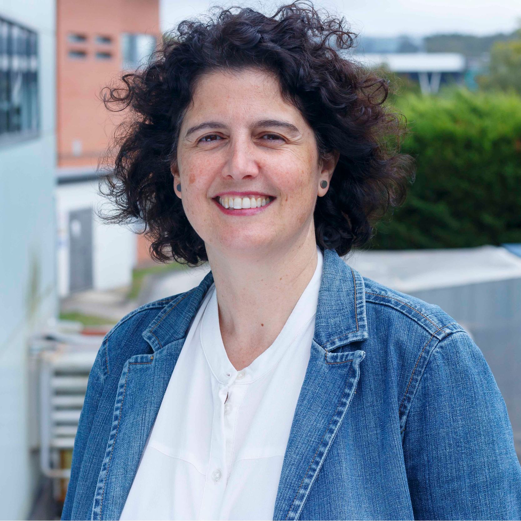 Mujer sonriente con cabello oscuro y chaqueta de jeans sobre una camisa blanca. Mujer sonriente con cabello oscuro y chaqueta de jeans sobre una camisa blanca.
