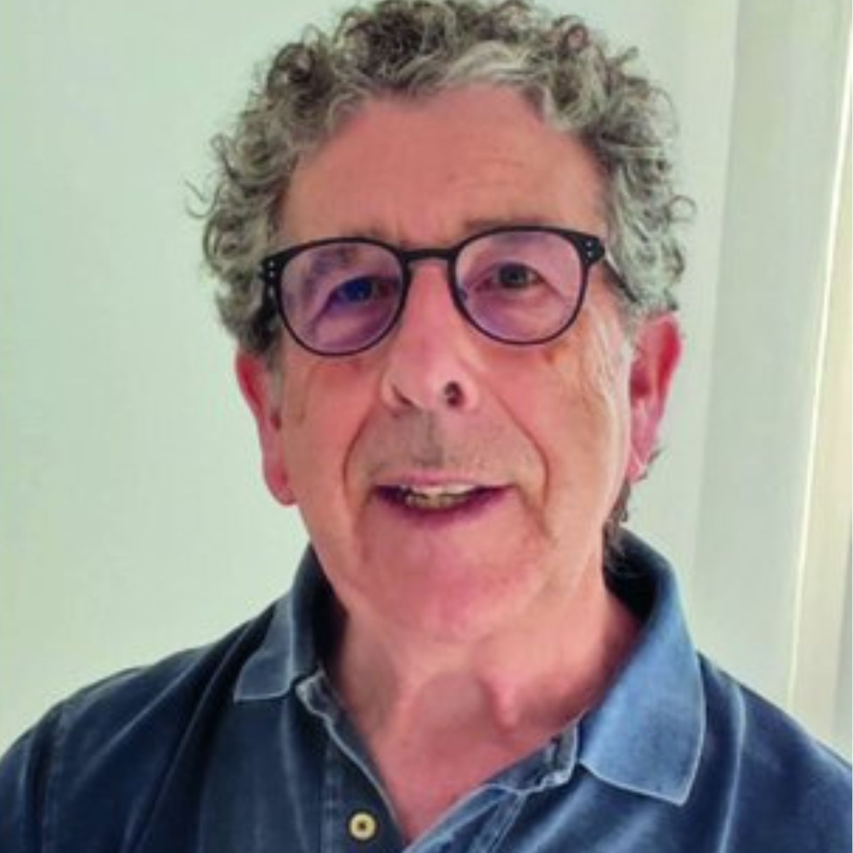 Varón con cabello rizado gris, gafas y camisa azul.