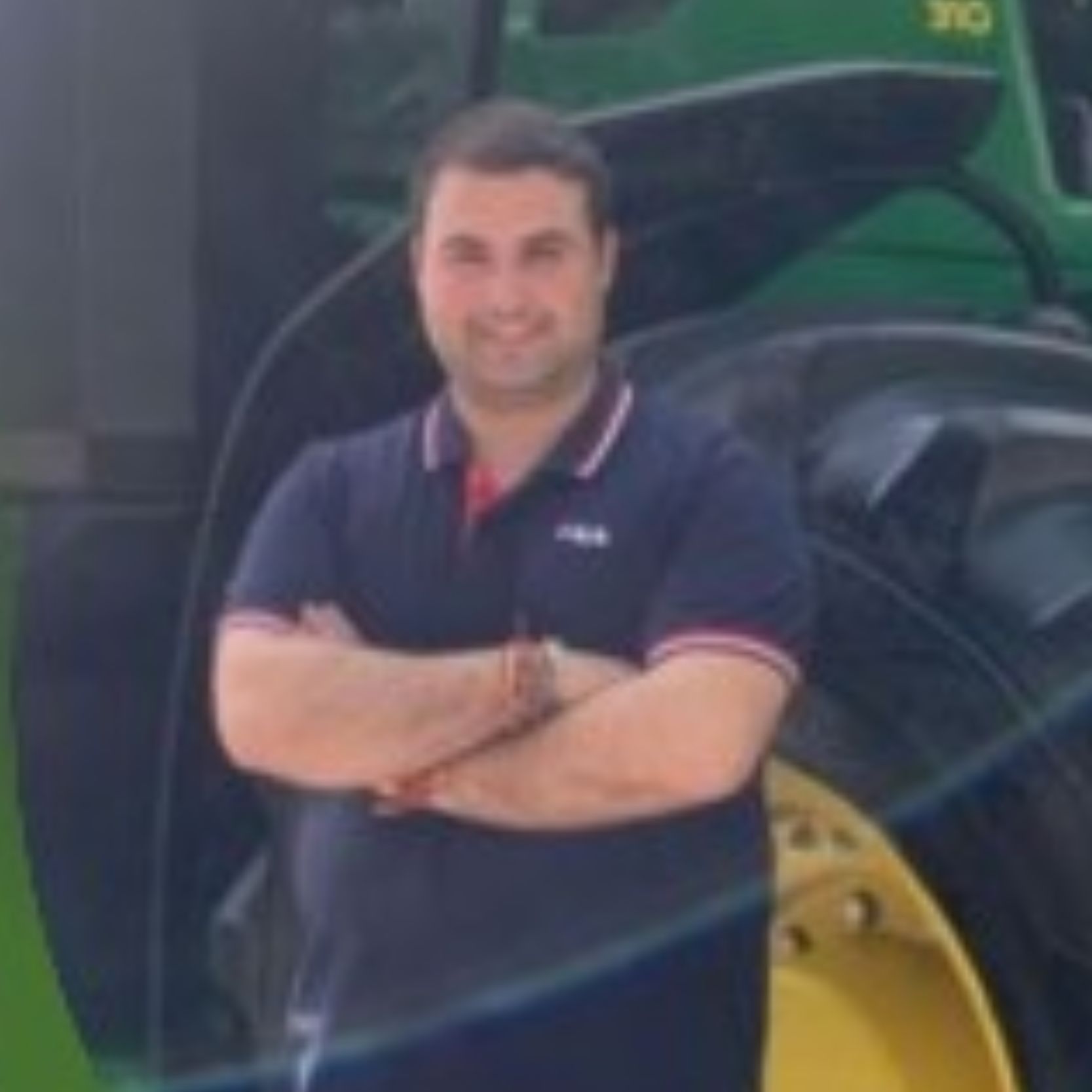 Ponente sonriente con polo azul marino y brazos cruzados, apoyado frente a un tractor agrícola verde y negro.