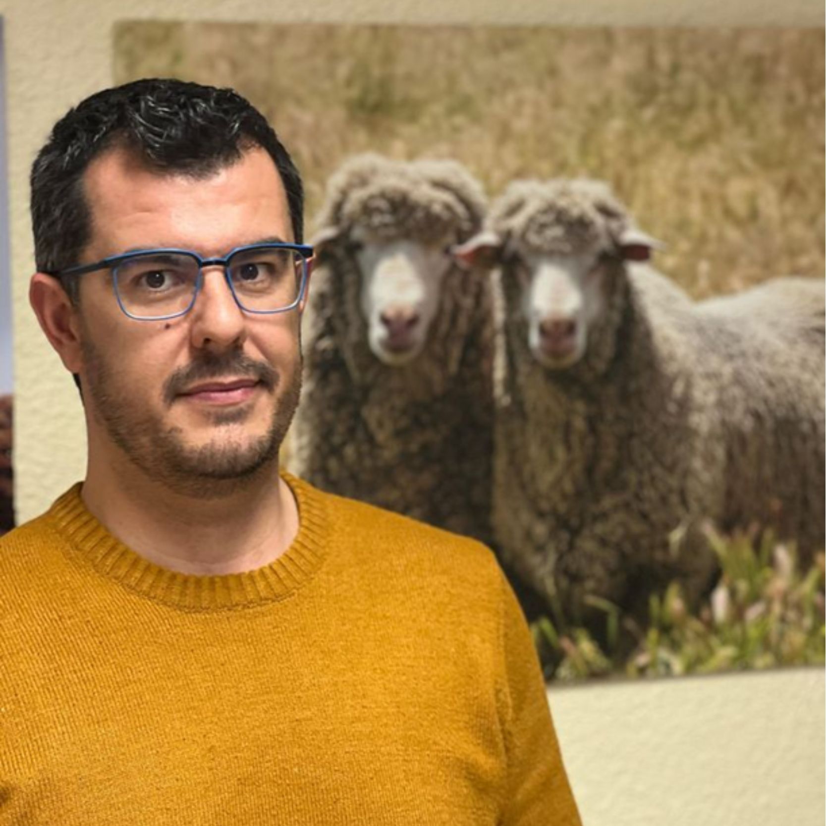 Un hombre sonriendo con gafas delante de un cuadro con dos ovejas.