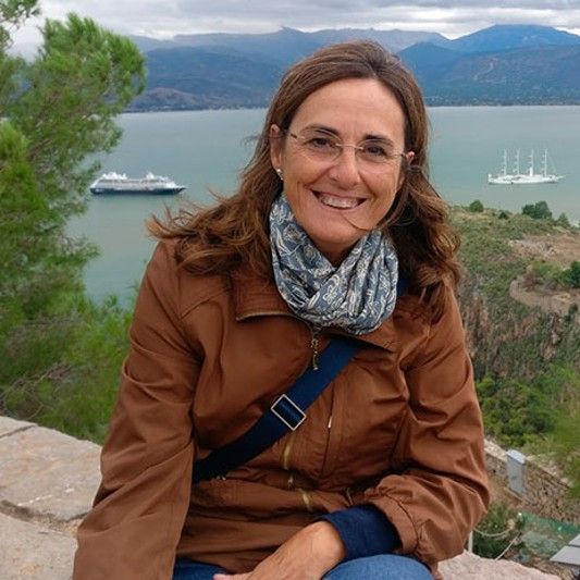 Mujer sonriente con bufanda y chaqueta marrón, mar en el fondo.