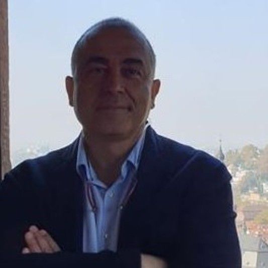 Hombre sonriendo con saco oscuro y camisa azul claro.
