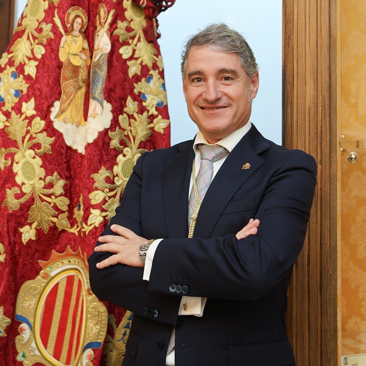 Hombre sonriente con traje formal frente a un tapiz histórico.