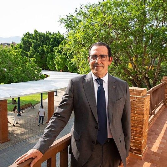 Hombre de traje posando en un balcón con un parque al fondo.
