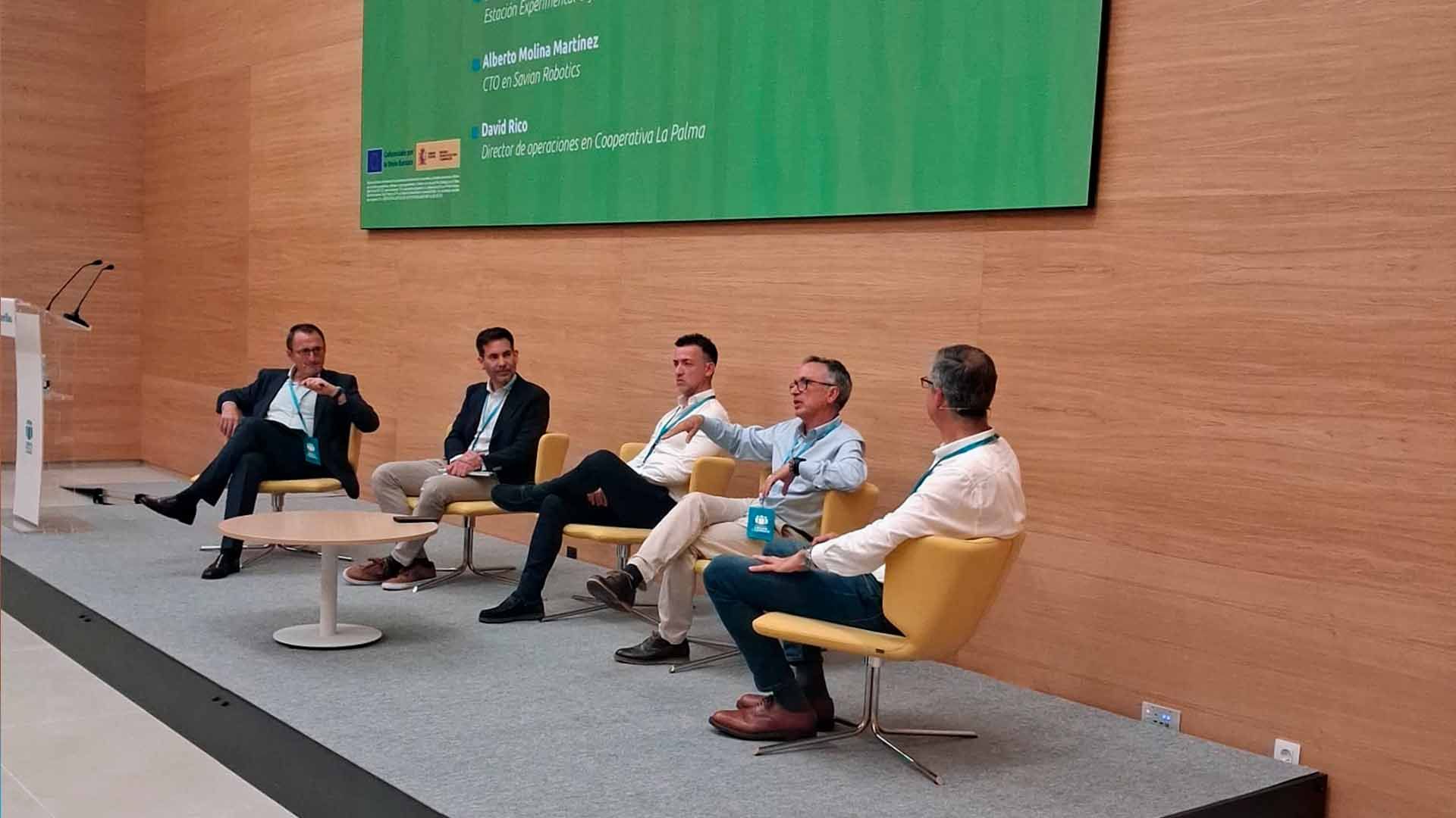 Cinco hombres sentados en sillas sobre un escenario con una pantalla grande al fondo.
