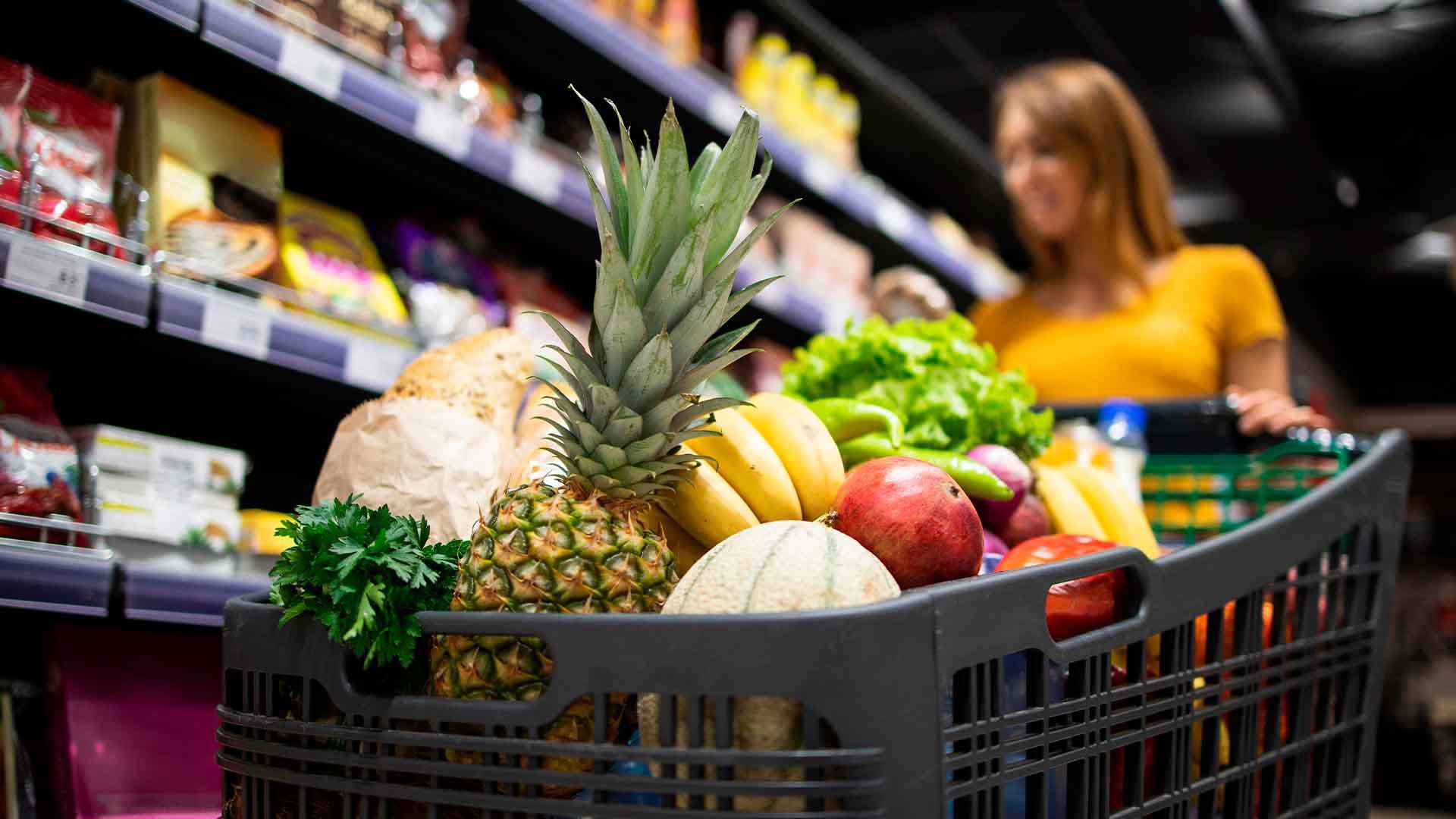 Mujer sonriente con un carrito de compras en un supermercado con productos frescos.