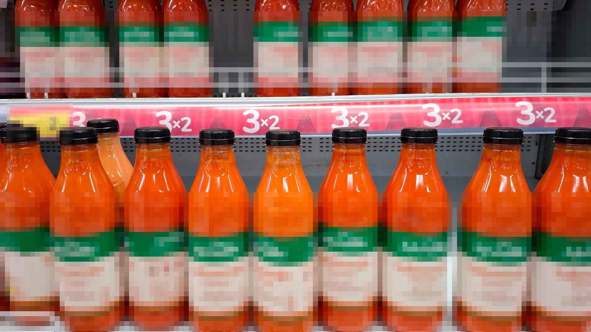 Múltiples botellas de bebida naranja en estante, con etiquetas borrosas y oferta 3x2.