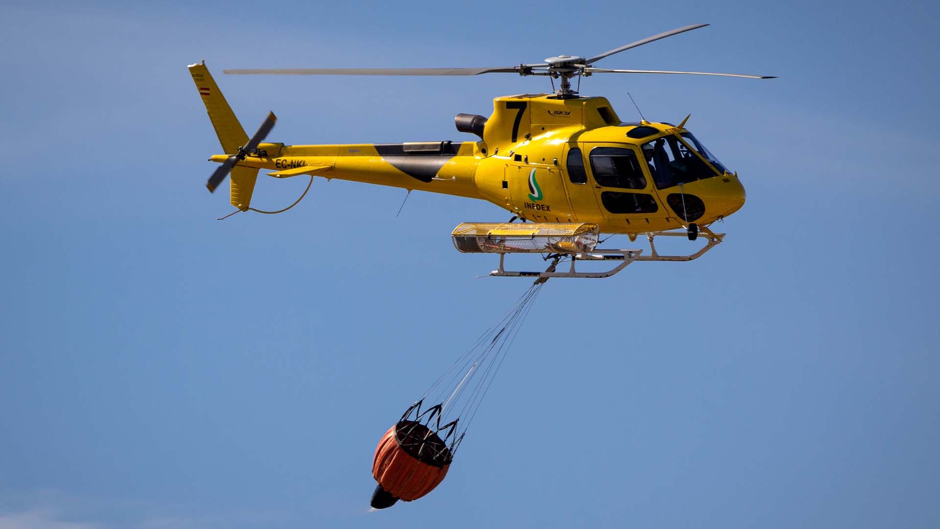 Un helicóptero amarillo en el aire con un balde naranja colgando con cables.