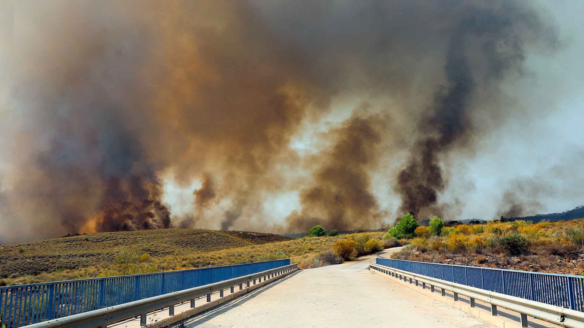 Humo denso y fuego de un incendio forestal cubren un paisaje árido con un puente.