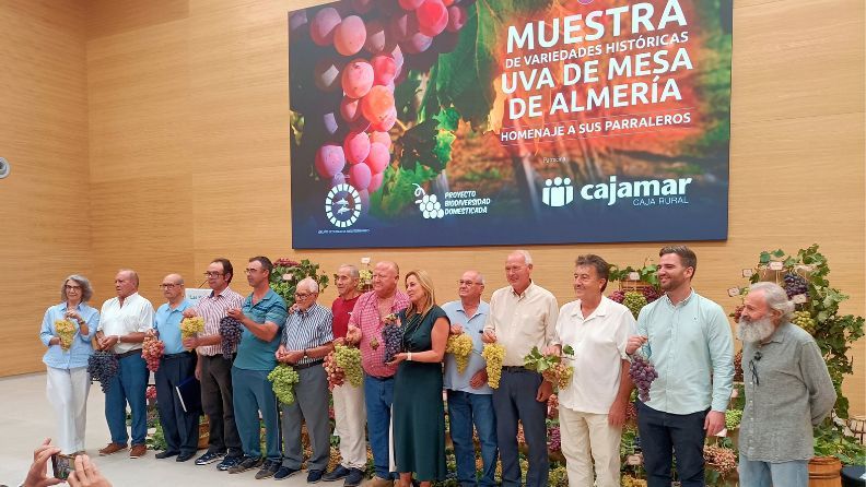 Grupo de parraleros junto a cartel evento sosteniendo uvas en la mano