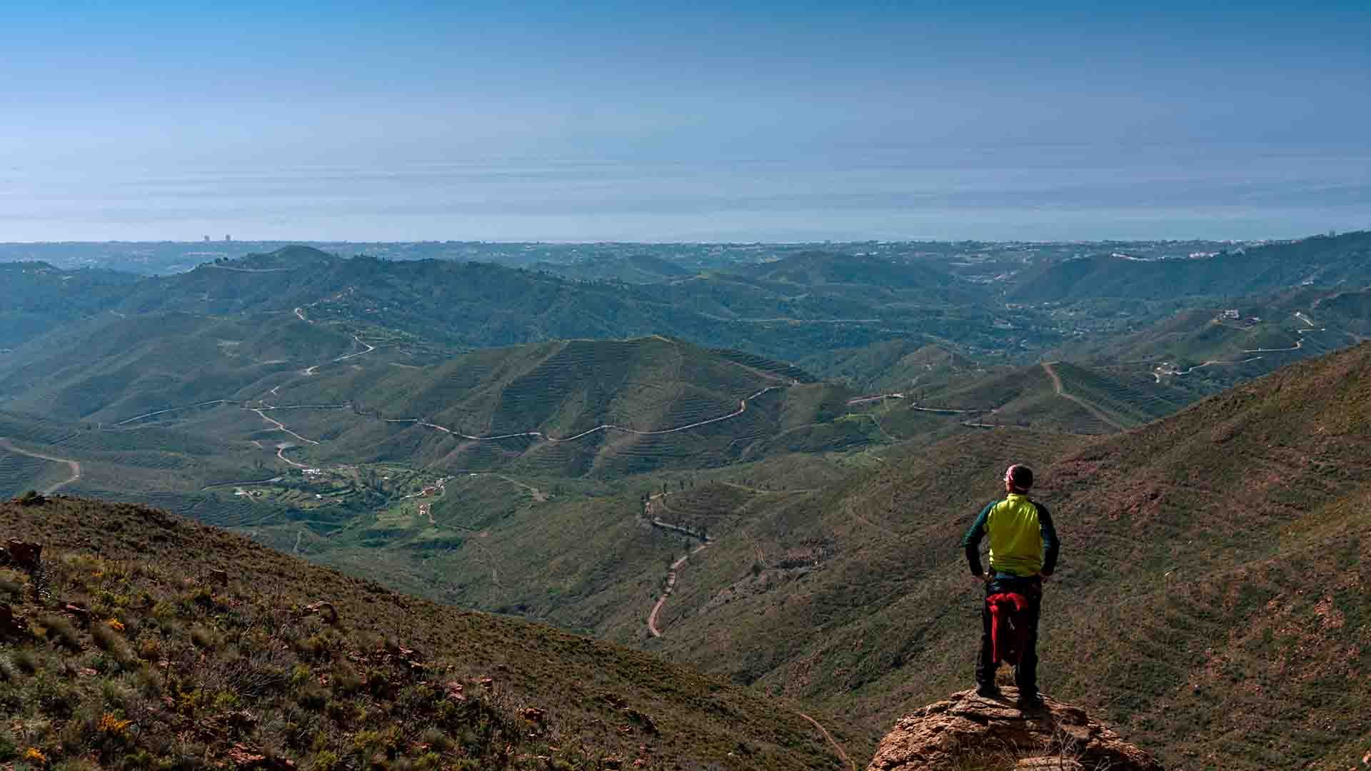 Caminante contempla valles y montañas verdes bajo cielo azul.